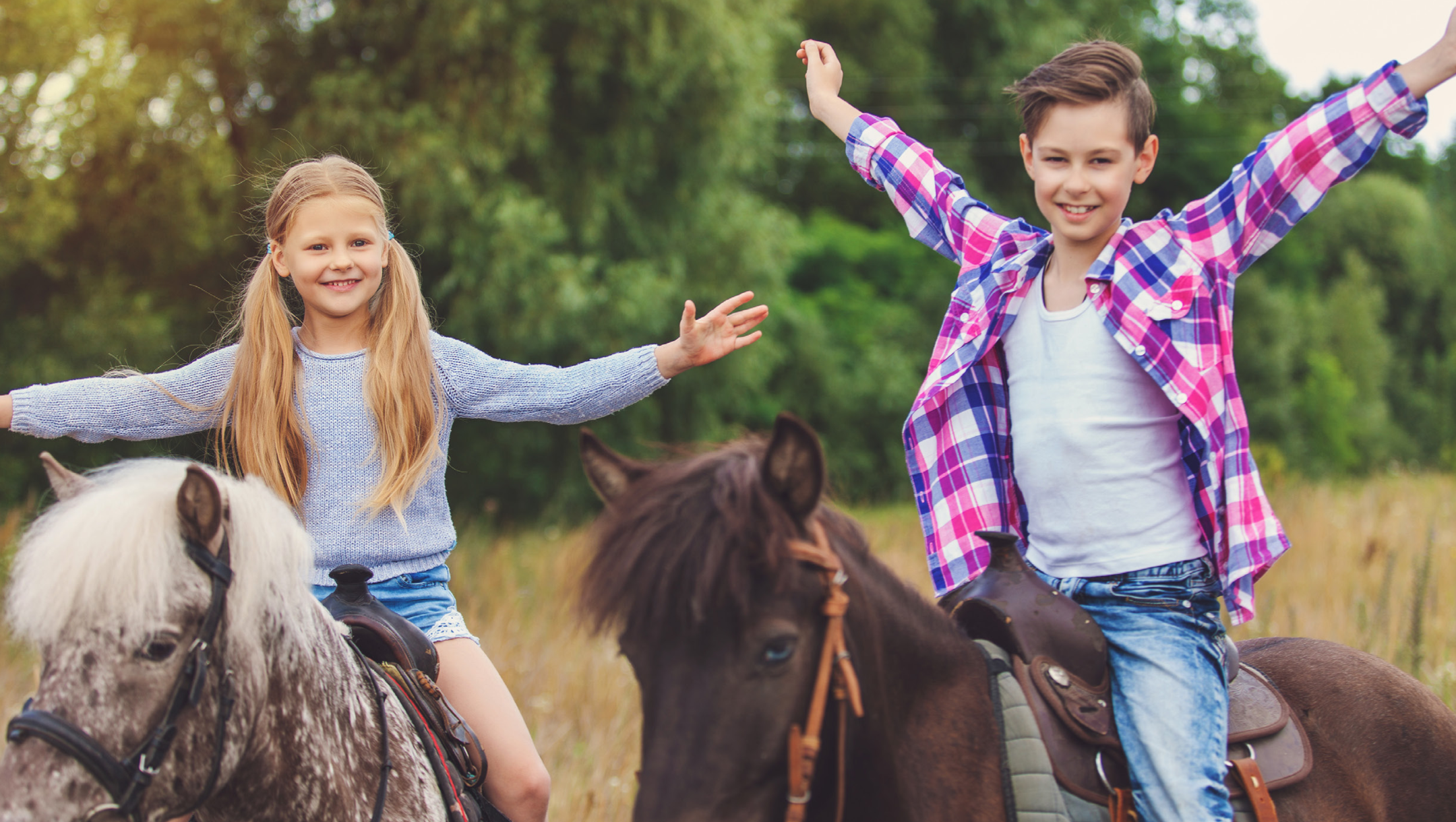 Two children looking happy with their hands raise in the air sitting on two horses