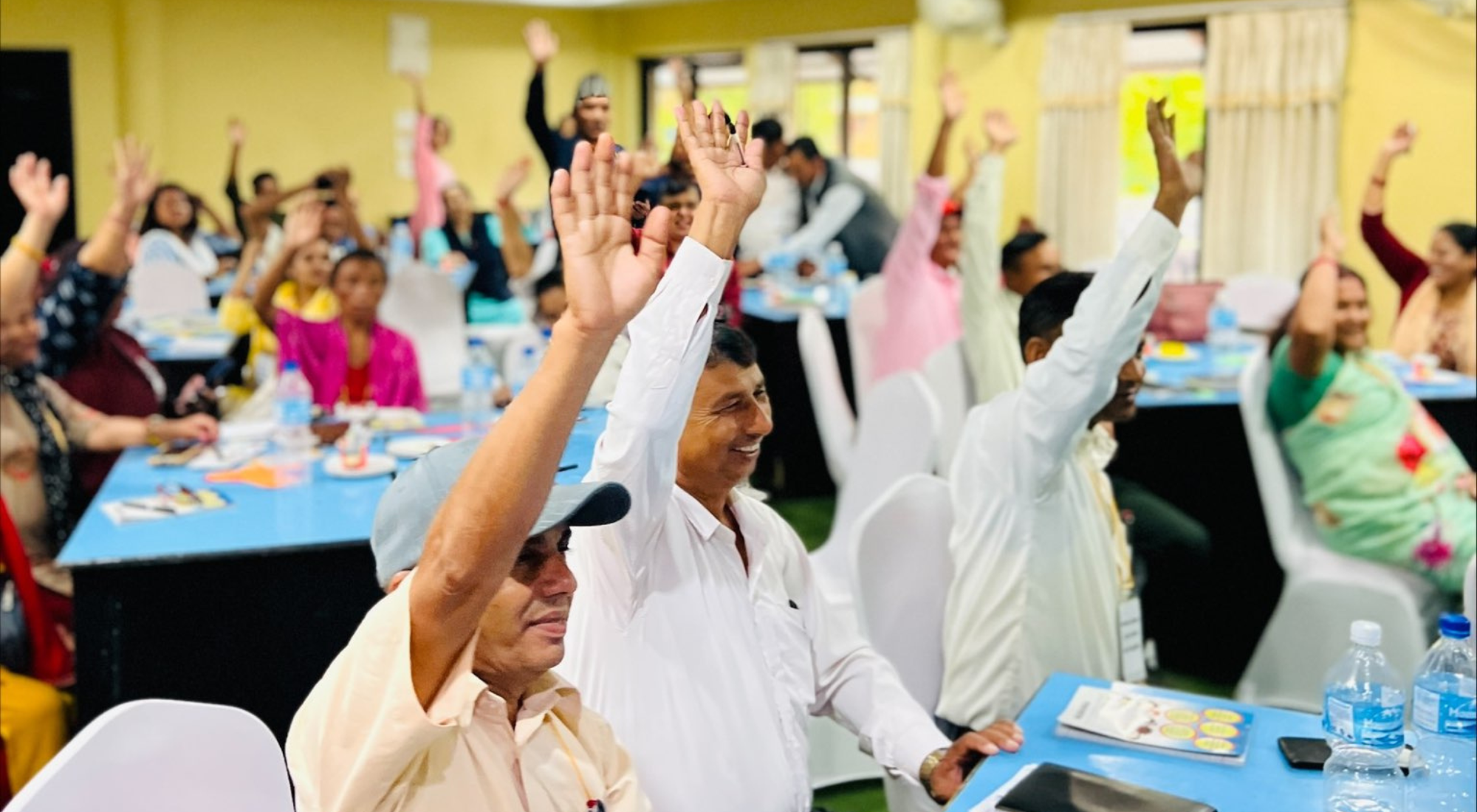 Members of organisations in Nepal attending a workshop. They are sitting and many are smiling with one hand raised.