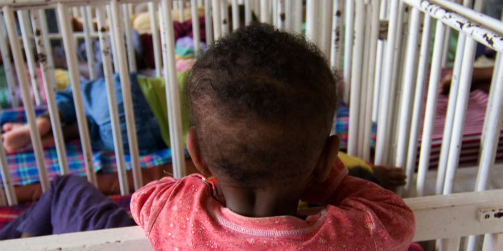 Child with back to the camera standing up in a crib in a room full of cribs