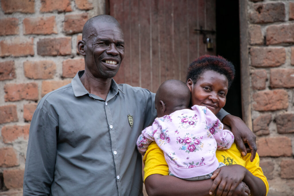 A Uganda couple who are foster carers looking at the camera and smiling holding the baby in their care