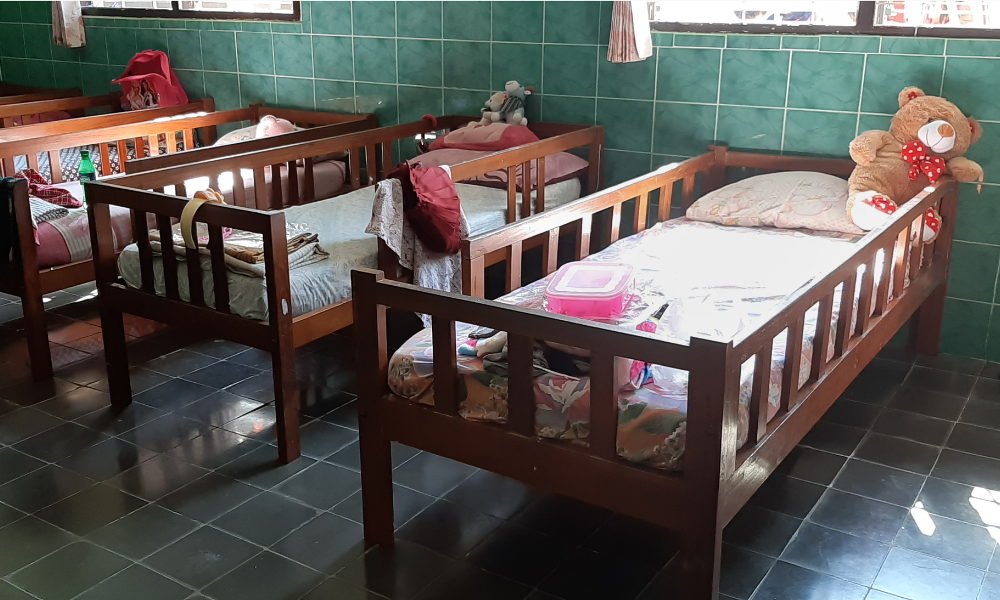 Cots lined against a wall in orphanage with teddy bear on one bed and light coming through windows.